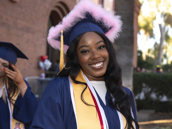 A young woman wearing a graduation cap decorated with pink feathers and a graduation gown smiles as she stands outside. 