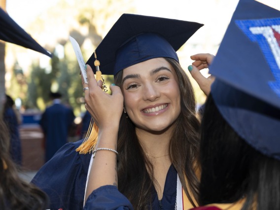 A smiling young woman wearing a graduation cap and gown has her hair adjusted by a friend. 