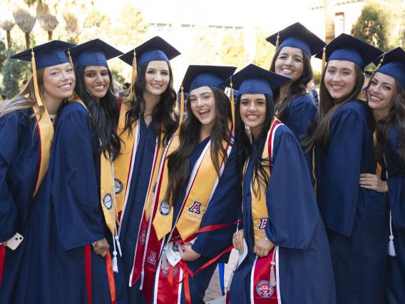 A groupl of eight young women stand together smiling, all dressed in graduation caps and gowns. 