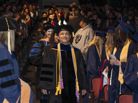 A line of nursing faculty walk down an aisle with students on both sides of them. All are wearing grauduation caps and gowns. 