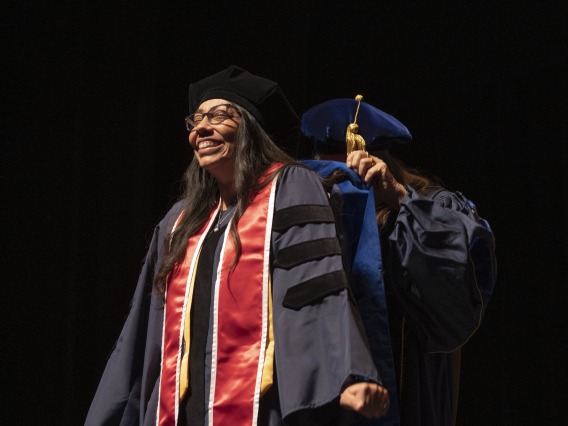 A nursing PhD candidate stands with a big smile on her face as a professor puts her graduation hood on her. Both are in caps and gowns. 