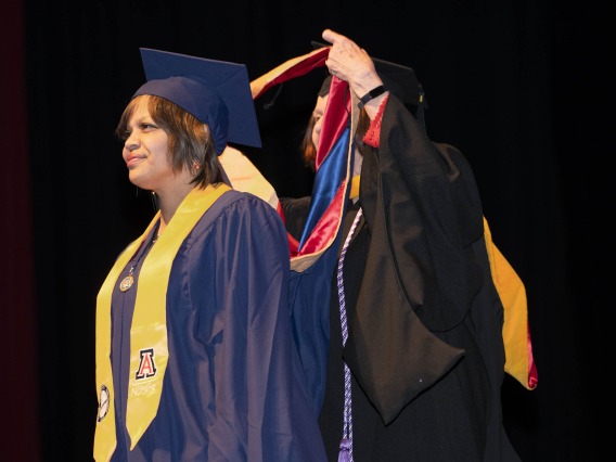 A female nursing student in graduation cap and gown stands looking forward as a professor places her graduation sash over her head. 
