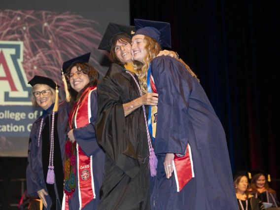 Two nursing students and two professors, all in graduation regalia, hug and smile on a stage. 