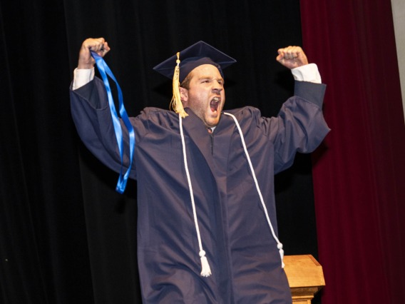 A young man wearing a graduation cap and gown raises both fists in the air and cheers. 