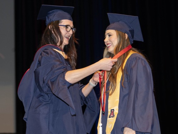 A nursing professor puts a lanyard on a nursing student. Both are wearing graduation caps and gowns. 