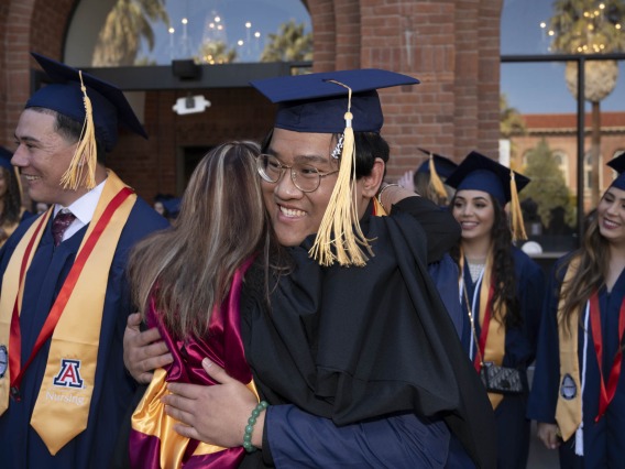 A young man in graduation cap and gown smiles as he hugs one of his professors outside. 