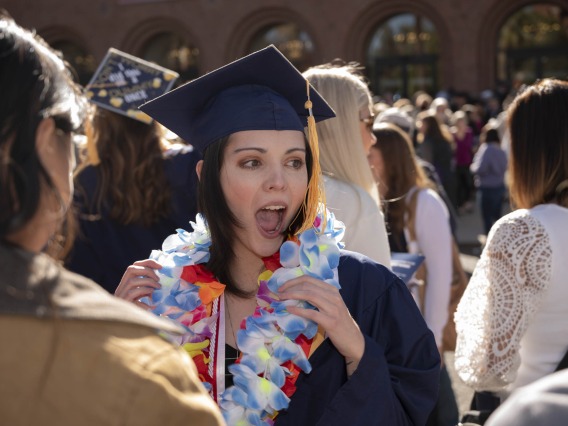 A young woman in a graduation cap and gown has her mouth open in a cheer in a crowd of people outside. 