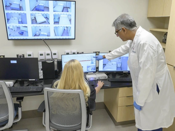 Man wearing white coat stands pointing at a screen. A woman with long blonde hair sits at a desk in front of the computer screen the man is pointing at.