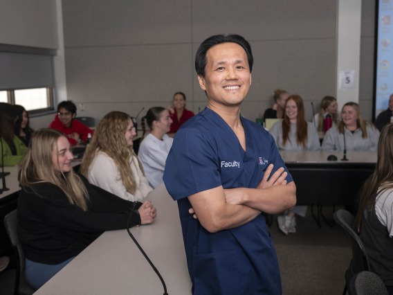 Portrait of Dean Hyochol Brian Ahn wearing navy blue nursing scrubs and standing in front of a class of nursing students.