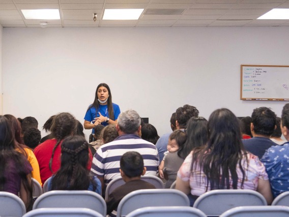 A woman in a blue shirt speaks to a crowd of migrants of all ages seated in metal folding chairs in a plain room.