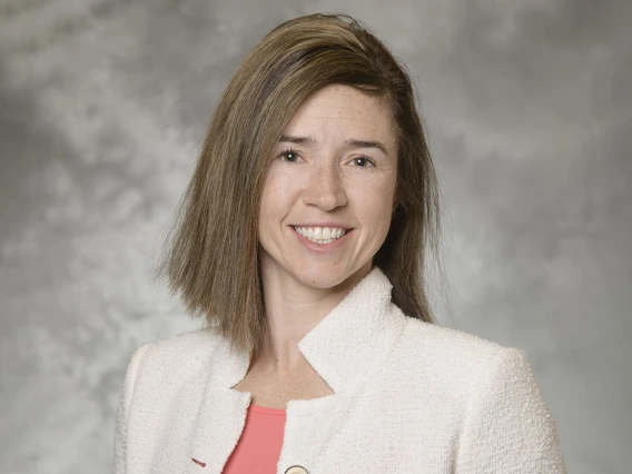 Portrait of a fair-skinned woman with soulder-length brown hair smiling. She is wearing a white blaser. 