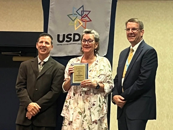 A smiling woman with gray hair holds a plaque as smiling men in dark suites stand on each side of her.