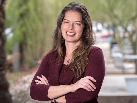 Portrait of a woman with long brown hair standing outside smiling with arms folded.