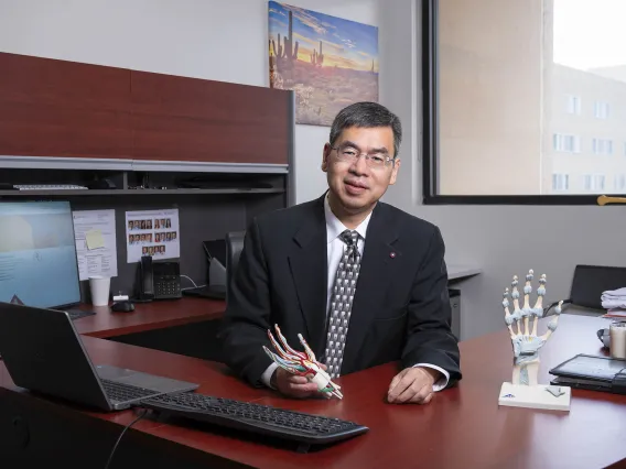 Zong-Ming Li sitting at his office desk holding an anatomical model of the inside of a hand. 