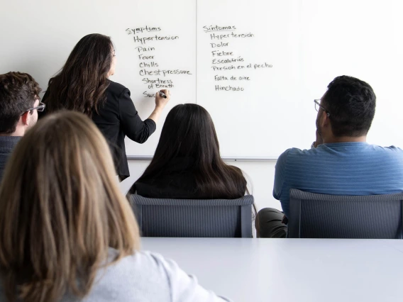 Students observe an instructor writing a list of medical terms in English and Spanish on a whiteboard