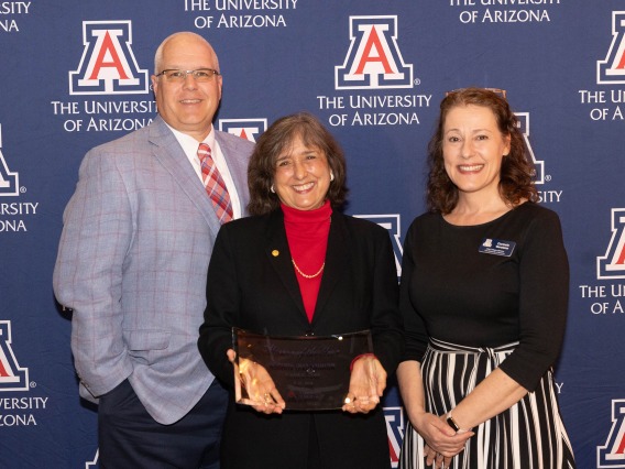 JP Roczniak, Roberta Diaz Brinton and Carmala Garzione stand in front of a backdrop with multiple University of Arizona logos. Brinton is holding a glass award. 