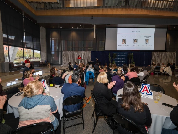 People sitting in a large room full of tables clap as they listen to a speaker at a podium and watch a projected slide on a screen in the distance. 