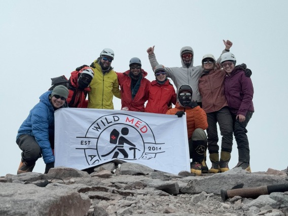 Nine people stand in front of a banner reading “Wild Med” at the top of Aconcagua.