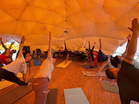 Inside a domed tent, a group of people on yoga mats lean on one arm and reach the other arm up toward the ceiling.