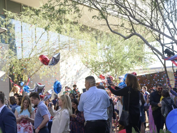 A crowd of people stands outside with confetti flying in the air. Lots of balloons decorate the space. 