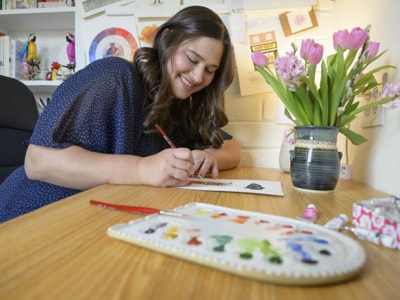 Anna Alkozei smiles as she holds a watercolor brush and paints a picture. A paint palette is in front of her and a vase of tulips in the background. 
