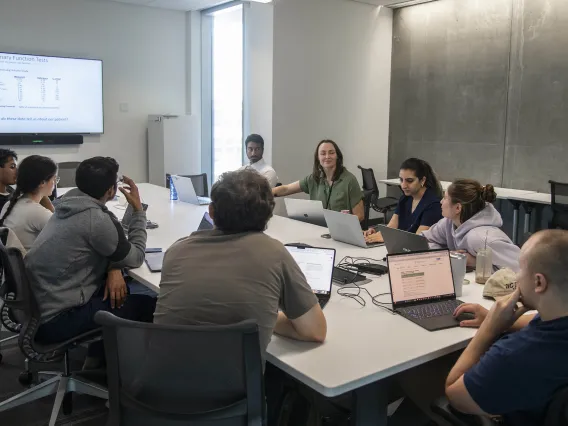 Halszka Glowacka sits at a conference table surrounded by students with a screen with writing on it in the background.