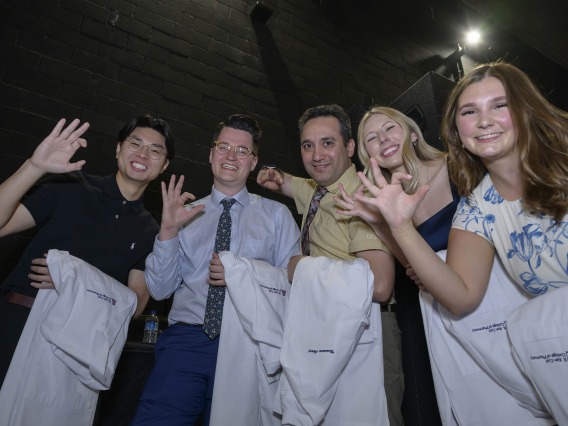 Five University of Arizona R. Ken Coit College of Pharmacy students give the Wildcat hand sign while waiting backstage with their white coats draped over their left arms. 
