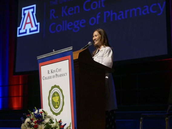 Professor Jeannie K. Lee speaks at a podium on a stage with the University of Arizona logo and “R. Ken Coit College of Pharmacy” projected on a screen behind her. 