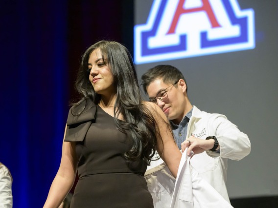 A student in a black dress and long dark hair puts on her pharmacy white coat with the help of a faculty member standing behind her.  