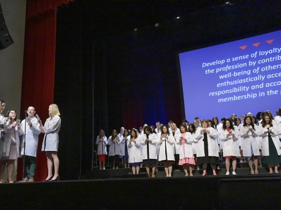 Four University of Arizona R. Ken Coit College of Pharmacy students stand around a microphone on the left while dozens stand in the background, all reading a professionalism pledge from cards they are holding.