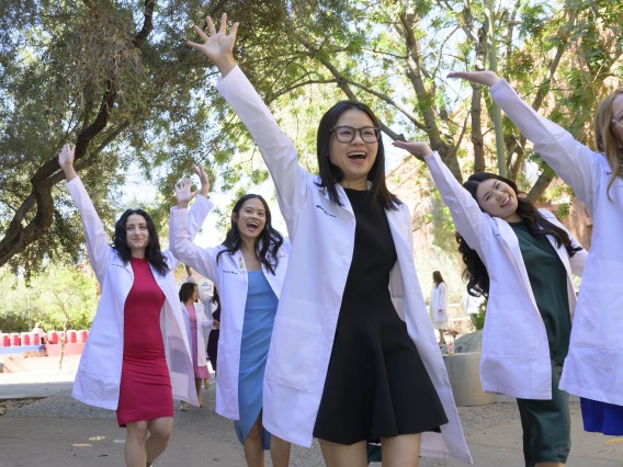 Several University of Arizona R. Ken Coit College of Pharmacy students wearing white coats raise their hands and cheer as they walk outside. 