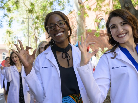Two UArizona R. Ken Coit College of Pharmacy students smile and make the Wildcat sign with their fingers as they walk outside after receiving their white coats at a ceremony.