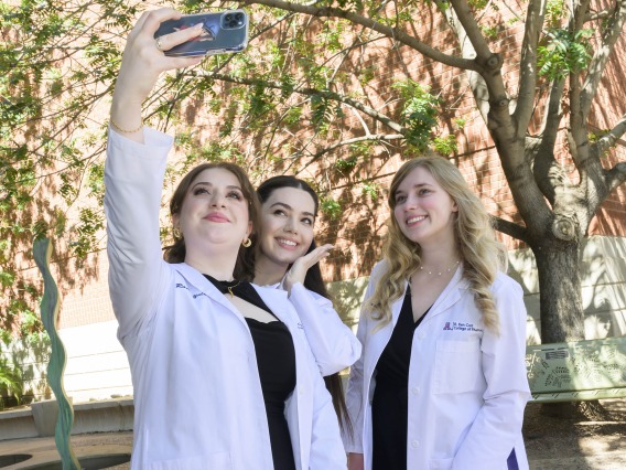 Three University of Arizona R. Ken Coit Pharmacy students in white coats pose for a selfie in an outdoor space. 