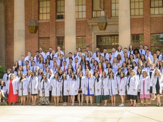 128 University of Arizona R. Ken Coit College of Pharmacy third-year students gather for a group photo on the steps outside a building wearing their white coats. 