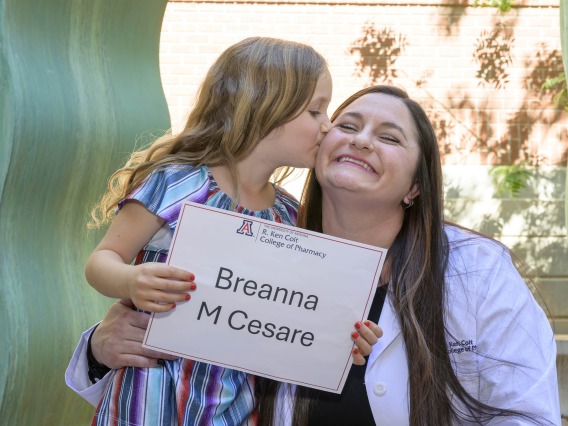 A young girl kisses her mom’s cheek while holding a sign that says “Breanna M. Cesare”. 