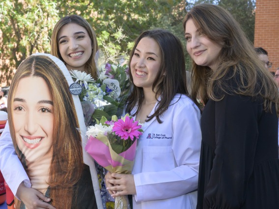 Two University of Arizona R. Ken Coit College of Pharmacy students and one graduate stand together with flowers and a large photo. 