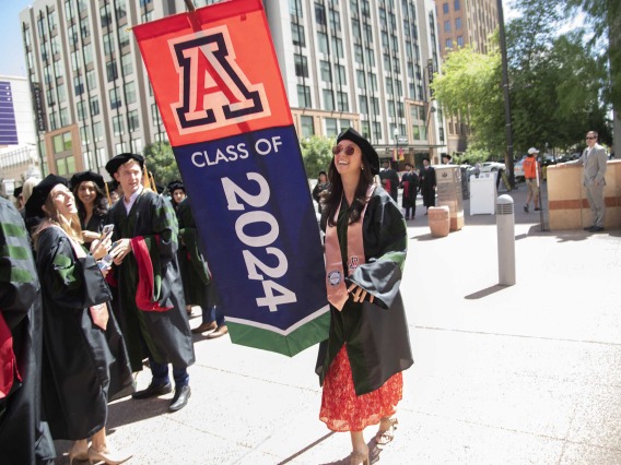 A University of Arizona College of Medicine – Phoenix student dressed in a graduation cap and gown carries a large banner that says “Class of 2024” as she walks outside. 