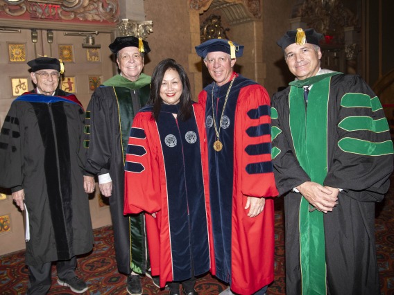 Five people dressed in graduation regalia stand together smiling. 