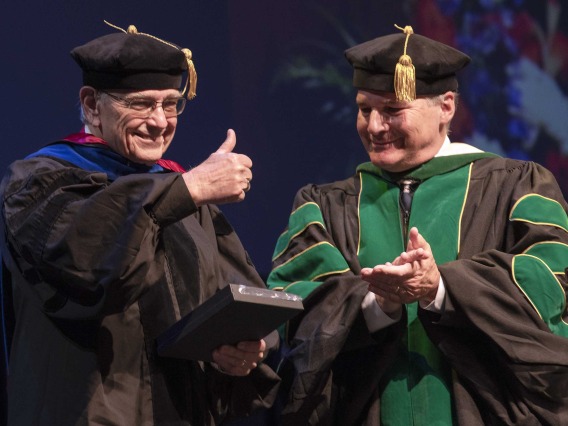 Two professors dressed in graduation regalia stand next to each other smiling as one hods a plaque and gives a thumbs-up. 