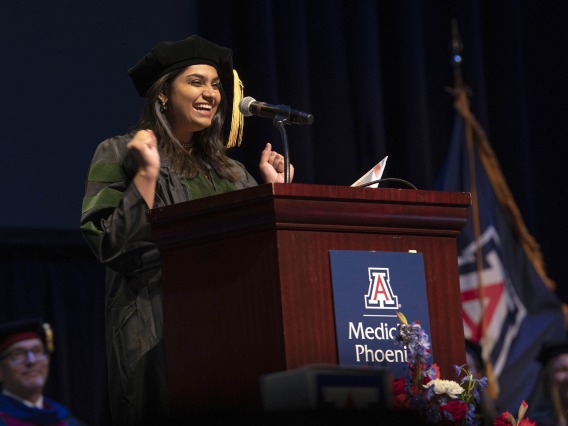 A University of Arizona College of Medicine – Phoenix student dressed in graduation regalia stands at a podium, smiling and gesturing with her hands, during the commencement ceremony.