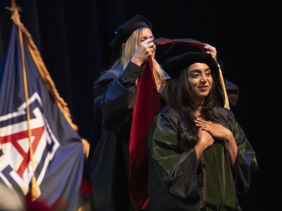 A University of Arizona College of Medicine – Phoenix student in graduation regalia holds her hands over her heart as a professor places a graduation hood on her.