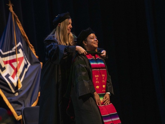 A University of Arizona College of Medicine – Phoenix student in graduation regalia smiles as a professor places a graduation hood on her.