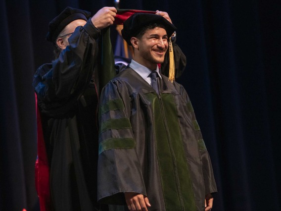 A University of Arizona College of Medicine – Phoenix student in graduation regalia smiles as a professor places a graduation hood on him. 