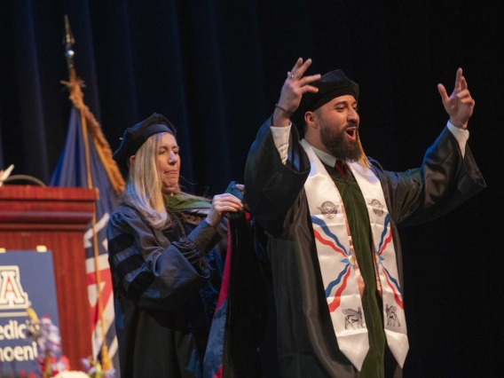 A University of Arizona College of Medicine – Phoenix student in graduation regalia holds his hands in the air in celebration as a professor standing behind him prepares to place a graduation hood on him.