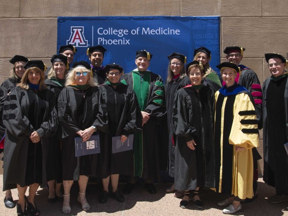 Over a dozen University of Arizona College of Medicine – Phoenix faculty members wearing graduation regalia stand in front of a sign that reads, “College of Medicine – Phoenix.”