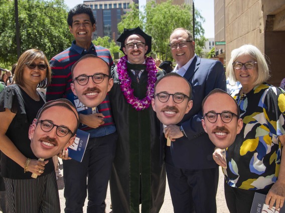 Several family members holding large faces on sticks surround a newly graduated medical student wearing a cap and gown after the University of Arizona College of Medicine – Phoenix commencement. 