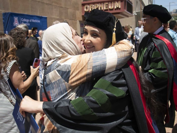 A woman in a hijab hugs her daughter, who is wearing graduation regalia outside amid a crowd of people.