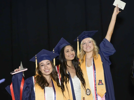 Three University of Arizona College of Nursing students in graduation regalia smile and raise hands in the air in celebration of their graduation.  