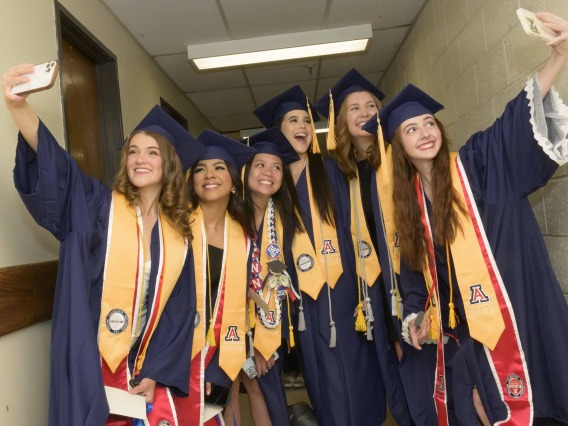 Six University of Arizona College of Nursing students dressed in graduation caps and gowns stand in a hallway while two of them are taking selfies. 