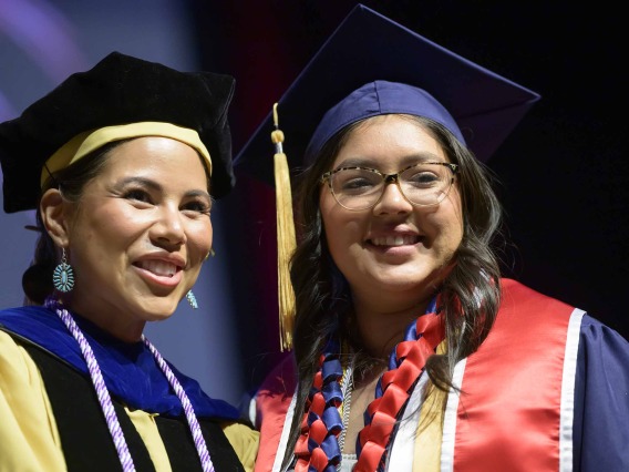 A University of Arizona College of Nursing professor and student, both wearing graduation caps and gowns, stand side by side smiling.